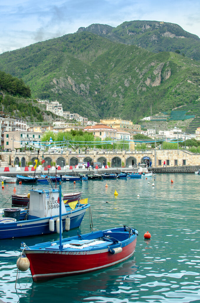 A picturesque view of colourful fishing boats in Maiori harbour, surrounded by scenic hills and the sea. The sea water is turquoise. The boat in the foreground is made of wood and is red, white, and blue.