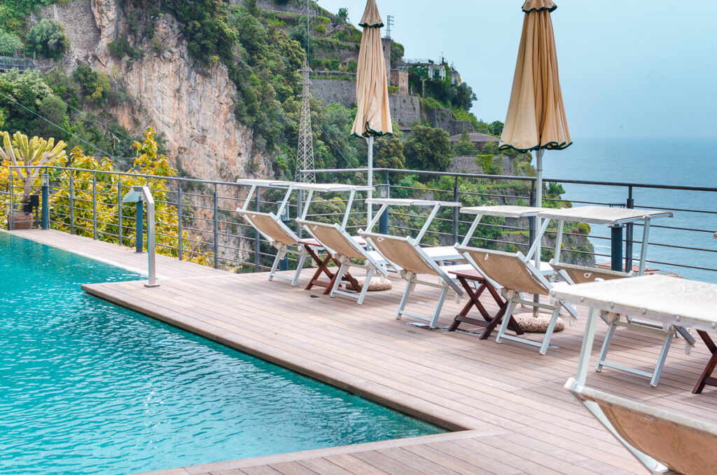 A photo of a beautiful swimming pool at the Hotel Botanico San Lazzaro in Maiori on the Amalfi coast, overlooking the sea. The pool is sparkling blue and inviting, with lounge chairs and umbrellas on the surrounding terrace. The image is used for illustrative purposes only and is not part of the presented list.