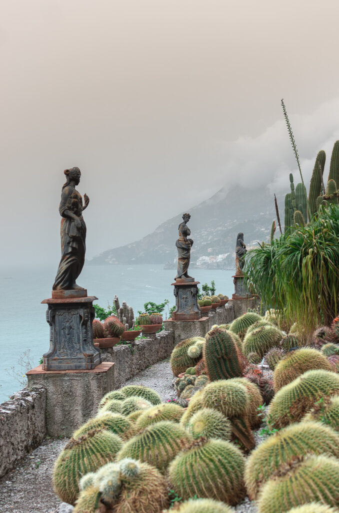 The view from the Botanical Gardens terrace in Maiori. The gardens are located at the Hotel Botanico San Lazzaro. In the picture, there is a terrace full of cactus plants, and neoclassical-style statues standing on the surrounding walls. The view includes the Amalfi coastline and the sea in the distance, which is almost completely covered in mist. The photo was taken on an overcast, rainy day.