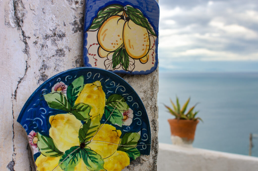 Detail of on old wall decorated with broken ceramic in Raito, Vietri Sul Mare, Italy. The wall has two ceramic plate on it, both hand painted with lemons. Blue and yellow, The background is white wall with aloe pat in a pot and the sea with clouds.