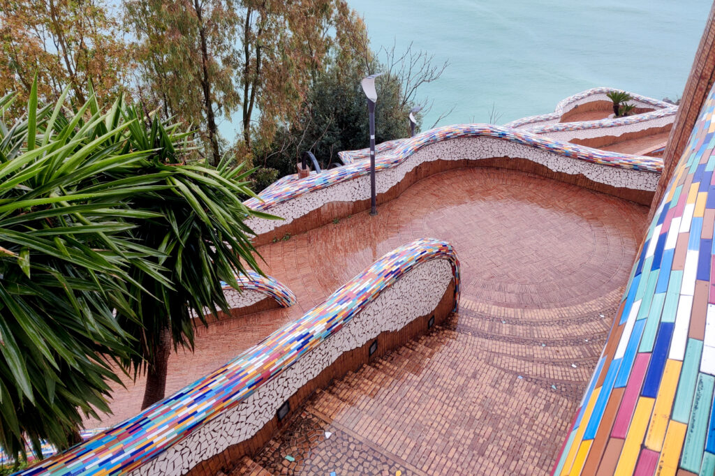 A photo taken from above of the garden at Villa Communale in Vietri sul Mare on a rainy day. The steps leading down through the garden are visible in the front of the photo. In the background, the sea can be seen through the rain. The walls are decorated with colourful and vibrant ceramic tiles. The surface of the ground is wet from the rain. Lush green vegetation and trees are on the side.