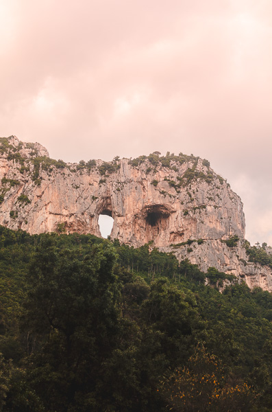 Fascinating natural rock formation in Positano, featuring a captivating hole in the rock wall. The sunset is casting a warm, orange glow on the rocks and clouds.
