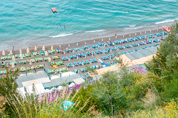 Aerial view of Fornillo Beach in Positano, one of the Amalfi Coast's top beaches. The beach is known for its tranquility and abundant free sun loungers. The loungers are blue and white striped. Idyllic spot with teal blue waters with some people enjoying a swim on this picture.