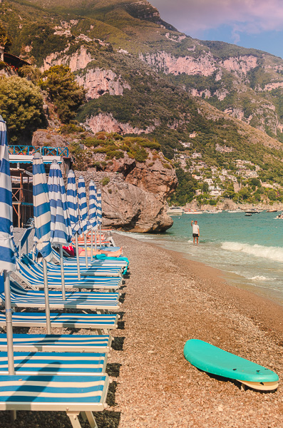 Rows of nearly empty sun loungers on the pristine Fornillo Beach in Positano.  The umbrellas are closed, and they are blue and white striped. A vibrant teal surfboard is left on the shoreline.  Almost the same color as the sea. In the distance, a lone man stands in the crystal-clear waters. The backdrop features the majestic cliffs and mountains of Positano.