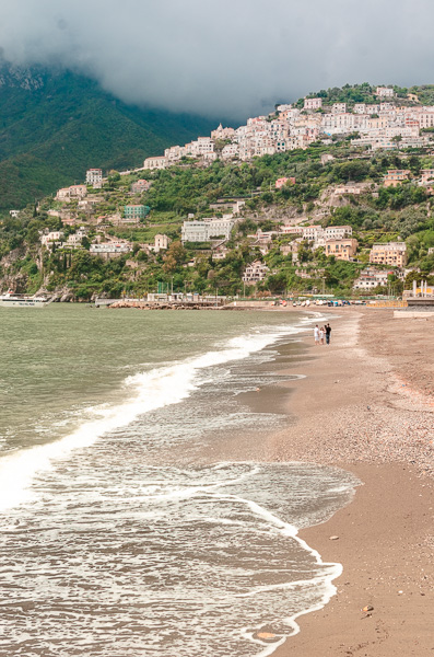 Vertical view of the beach of Vietri Sul Mare. On the Amalfi Coast, Italy. The image captures gentle waves lapping at the shore, with distant beachgoers walking along the sandy coastline. In the backdrop: misty mountains and white houses atop, creating a breathtaking coastal scene.