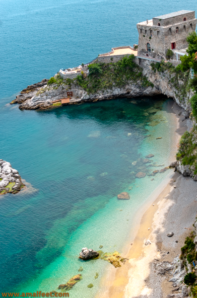 Aerial view of the Cauco Beach, near Erchie, Amalfi Coast. Cauco is one of the best beaches on the Amalfi Coast. This picturesque scene features a sandy shoreline merging with pebbles. The shoe has a golden hue. The bay is between rugged rock formations and a headland. The cape has an ancient tower on the top. Shallow and clear sea water transitions from deep blue to turquoise and teal. Beneath towering cliffs, lush vegetation thrives. The protected nature of this shore and the scenery makes it one of the finest Amalfi Coast beaches.