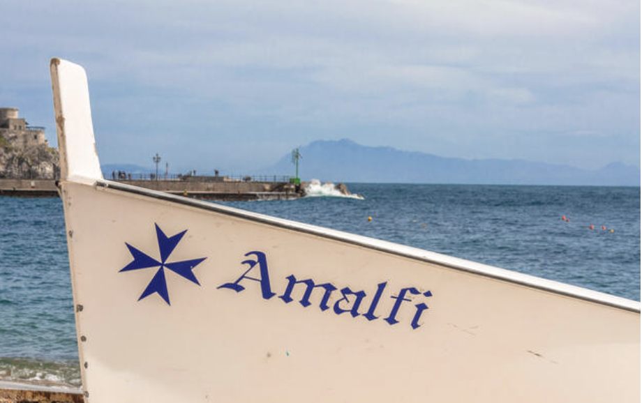 Boat at the town of Amalfi with text Amalfi and symbol of Amalfi. Image to illustrate Amalfi Capri ferry section of the post.