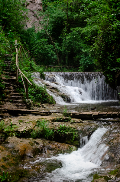 Small waterfall, and water stream on the foreground. Pathway with steps near the waterfall. Small wooden bridge above the river. Typical scenery of the Ferriere Valley