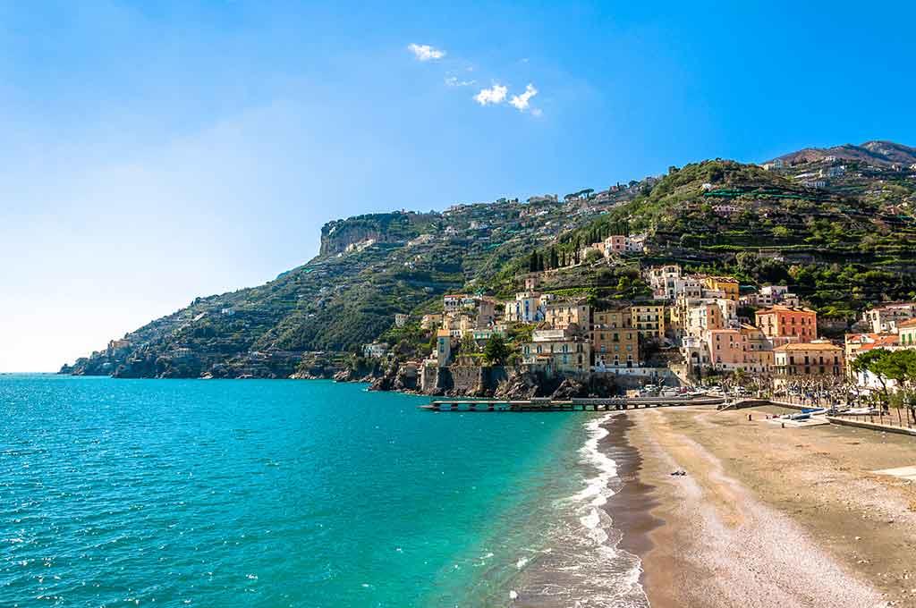 View of Minori Beach on the Amalfi Coast, Italy. In the foreground, the turquoise blue sea gently meets the sandy shore. In the background, a part of the village of Minori on the hillside, its houses. Beyond, the coastal landscape extends, with the iconic cliffs of Ravello crowning the mountain's peak, making it one of the best beaches on the Amalfi Coast.