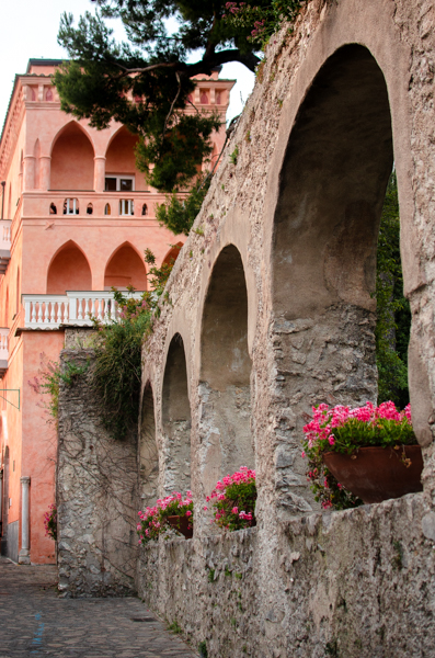 Ancient-looking arched stone walls along the small alleyway in Ravello. Pink flowers are growing in each arched opening. In the background the famous building of Palazzo Avino. The villa is painted pink and has balconies with pointed arches, and white balustrades.
