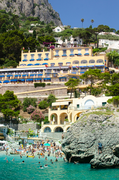 The Hotel Weber Ambassador as seen from Marina Piccola, Capri.
People are bathing in the sea in the foreground. The hotel is on the Cliffside. The hotel is yellow with blue sun covers above the windows. 