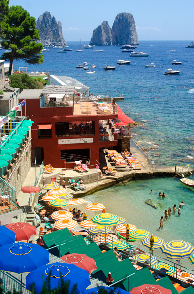 The seaside of Capri: striped umbrellas and a beach club, with the Faraglioni rocks in the background. Near Marina Piccola