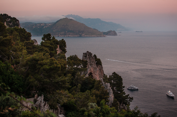 Sunset view from Capri, with Punta Campanella and the Amalfi Coastline visible in the distance. In the foreground, you'll see trees and rock formations, while two boats are anchored in the water.
