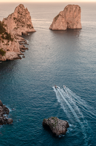 Two boats are heading towards the Faraglioni rocks at sunset. The boats are making a white trail left behind in the dark blue sea. 
