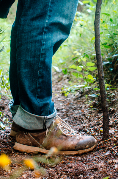 Hiker standing on the trail with a hiking stick. Close up on his feet. He is wearing blue jeans, and  brown hiking boots. The forest floor appears to wet and a bit muddy. Yellow flowers out of focus in the foreground.