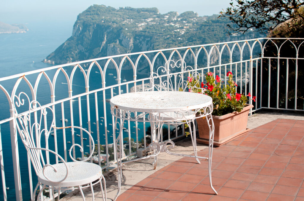 The panoramic terrace of Hotel San Michele in Capri. White fence, with a white table and chair, on terracotta terrace. Near the table there are red flowers in a pot. The Island of Capri and the Sea behind.