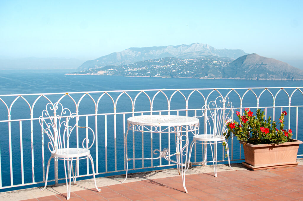 The panoramic terrace of Hotel San Michele in Capri. White fence, with a white table and chair, on terracotta terrace. Near the table there are red flowers in a pot. Mainland Italy and Sea behind.