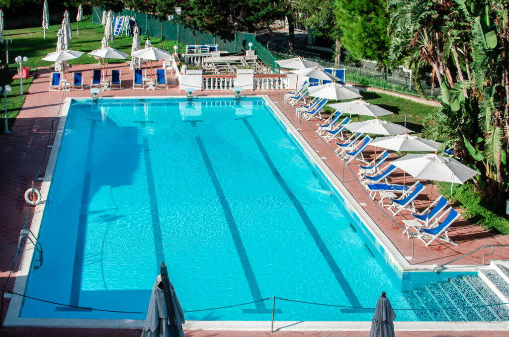 The large swimming pool of the Hotel San Michele, Capri, Italy.
Turquoise water in a semi olympic swimming pool, and a row of blue and white sun chairs at the poolside.
