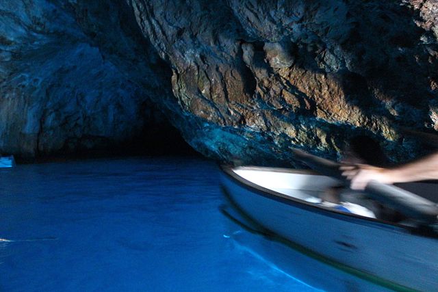 The prow of a rowboat gliding over glowing blue water, set against the rugged backdrop of a rocky cavern in the Blue Grotto, Capri.