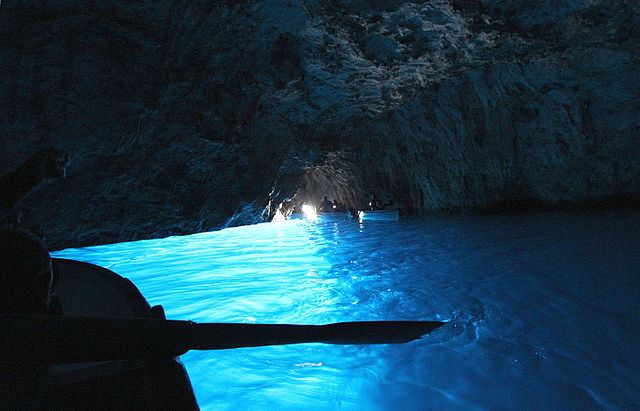 Silhouette of a rowboat's nose against the backdrop of glowing blue water inside the dark cavern of the Blue Grotto, an iconic natural wonder of Capri, Italy.