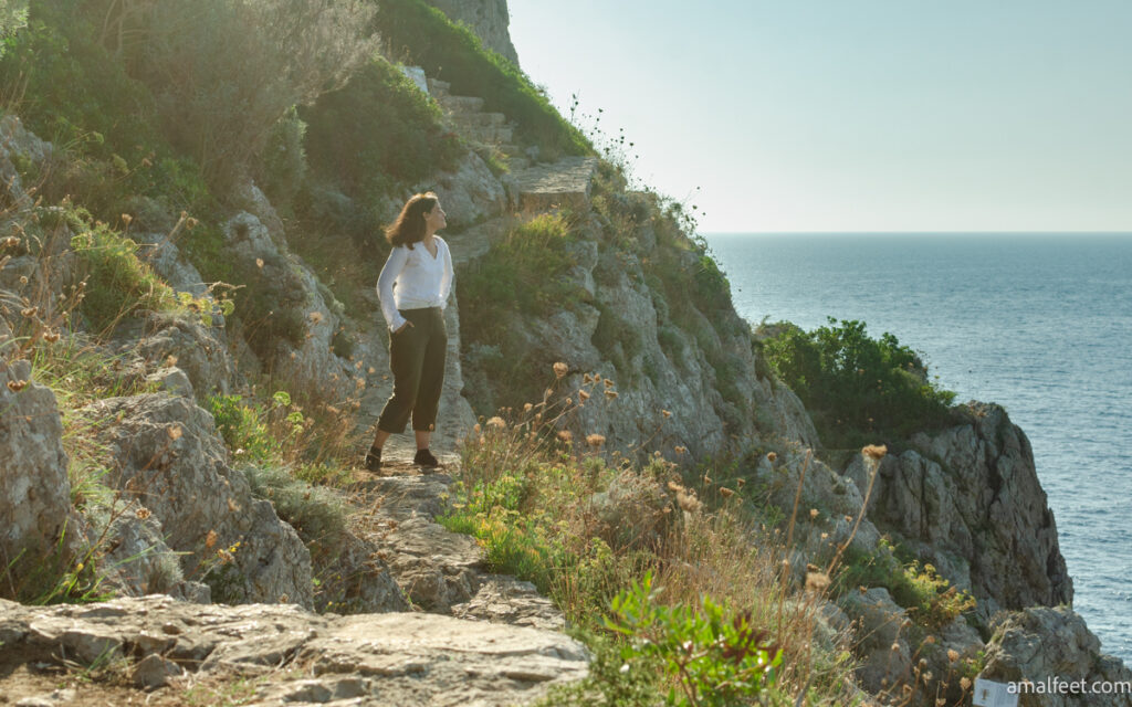 Me enjoying the sea breeze and the scenic view, during a hike in Anacapri.
Photo taken on the Sentiero dei Fortini. Girl wearing white shirt and khaki linen pants, standing on a paved footpath, on the cliffs. Looking to the direction of the sea.
