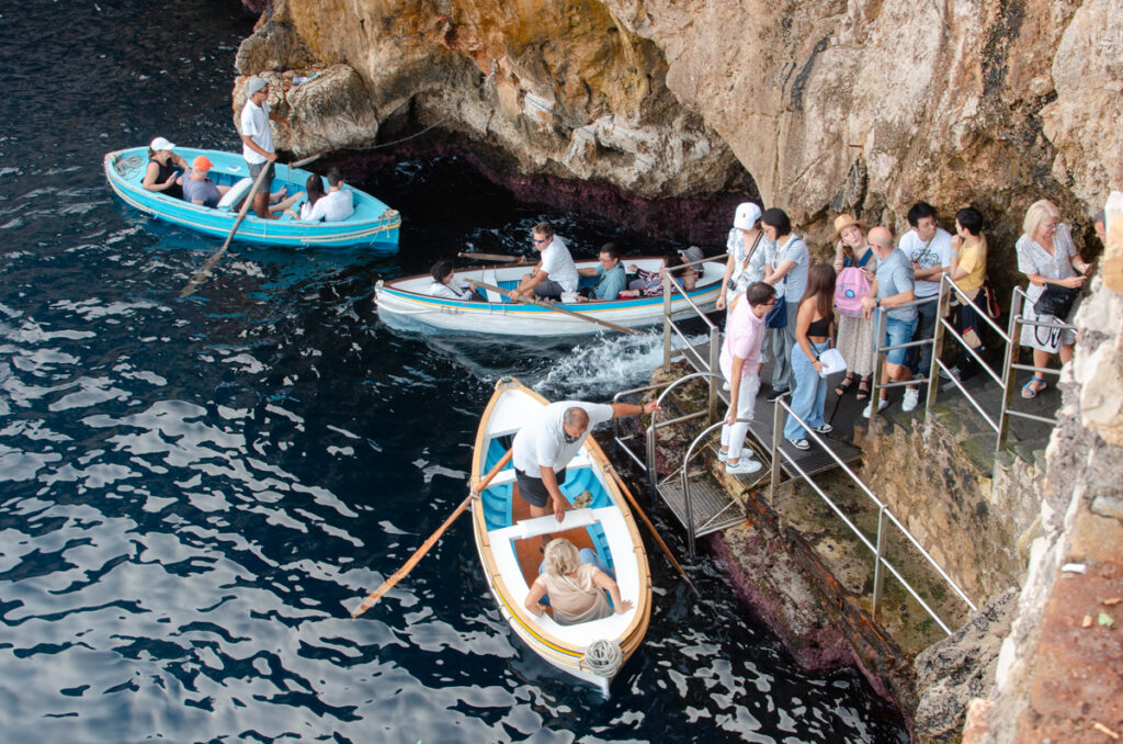 Action-packed scene at the Blue Grotto entrance: a rowboat emerges from the cavern's opening, another waits to enter, while a third picks up tourists from the shore. Visitors queue patiently on the steps to visit the stunning Blue Grotto.