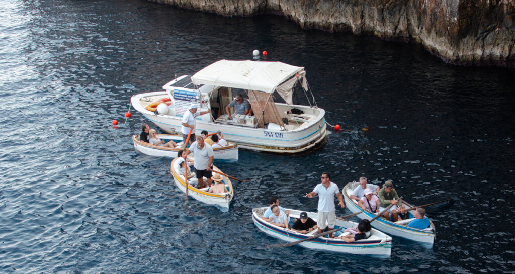 A scene near the entrance of the Blue Grotto, Capri. A larger boat, the floating ticket office, and four rowboats with boatmen and tourists aboard around it. Sea and rocky shores.