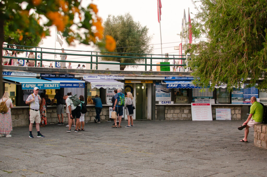 People waiting in lines to buy the ferry ticket in front of several small ticket offices. Cable stones on the square and flowers in the side of the image.