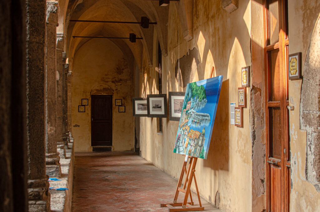 Inside of the Cloister. Ancient yellow walls and pointed arches. A colourful painting of the Amalfi Coast exhibited on the side, and several black and white images are hanging from the wall.