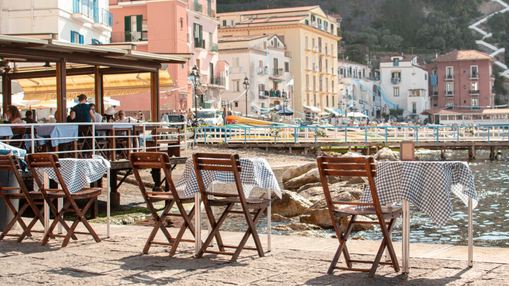 View of Marina Grande in Sorrento. Little tables with chairs near them facing the water. Pastel-colored houses and steep cliffs in the background.