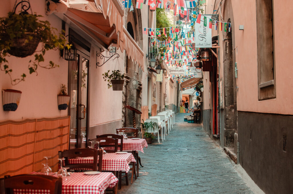One of the alleyways of Sorrento. Roadside restaurant tables, with classic red checkered tablecloths, cobblestone streets. Many little flags hang from the top of the street. 