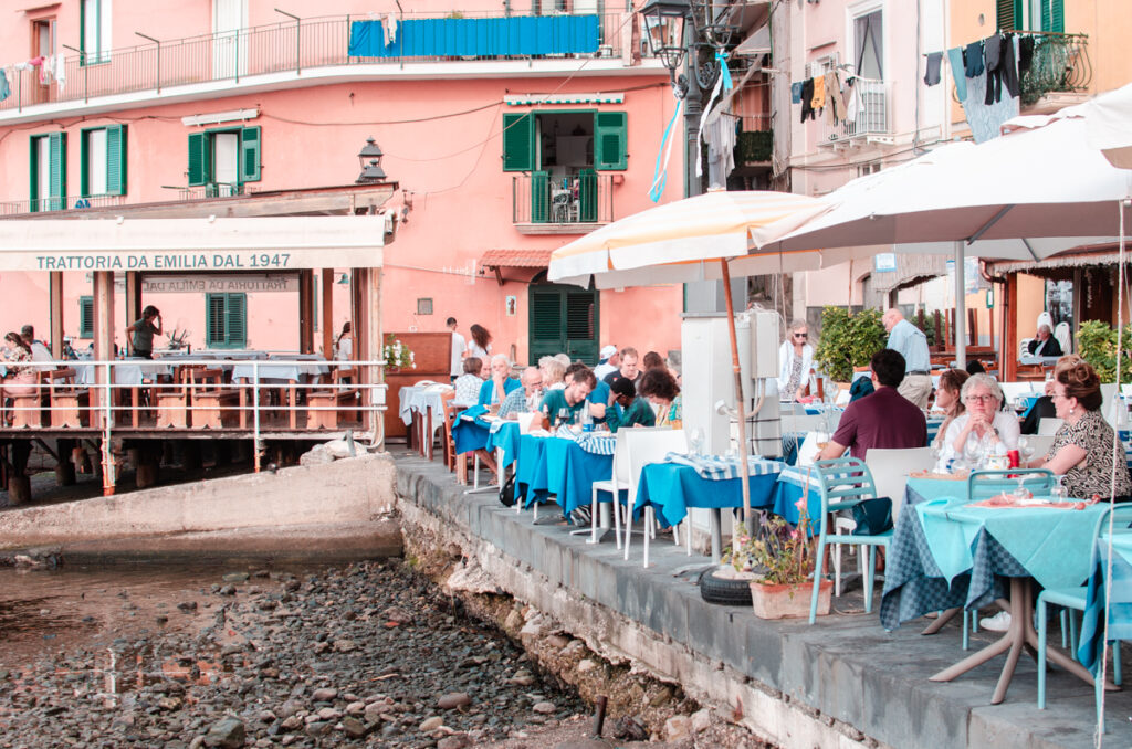 People eating by the seaside, small tables with blue tablecloths at Marina Grande, Sorrento. Pastel pink houses in the background.
