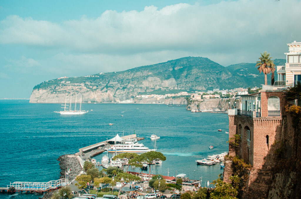 The view of the dramatic cliff from Sorrento, with the sea and the Marina Piccola ferry port in the foreground. A white sailboat is sailing on the water.