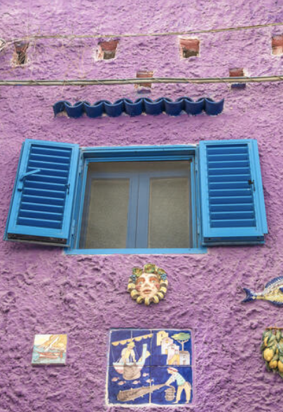 FFacade of the famous colourful houses in Vietri Marina. This is a deatil of the purplle wall with bright bllue window blinds open. many ceramic objectt incorporated in the wall. 