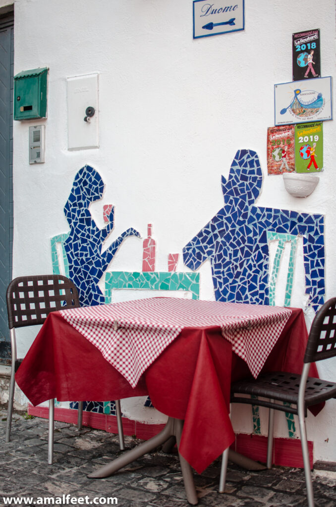 Funny cerammic decorationns at the outside of the local restaurant lucia 34. Behind a real table of the restaurant, there is a mosaic on the wall. the mosaic feauters a silhutee off a couple enjoying dinner with a bottle of wine.