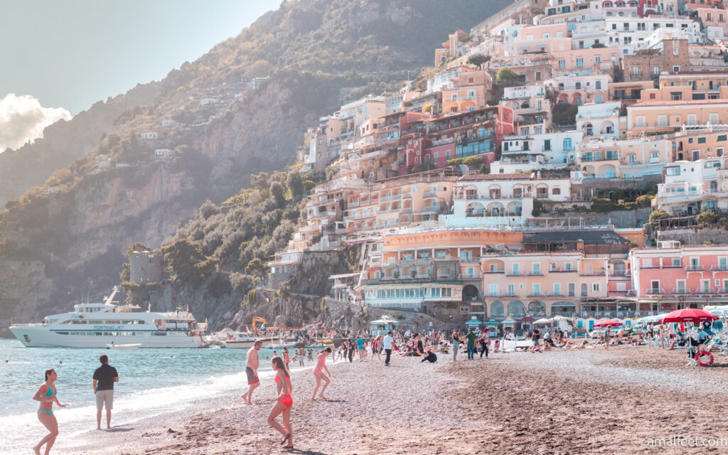 People enjoying a sunny day on the main beach of Positano. Behind the towering cliff, fully built in with pastel colured houses.