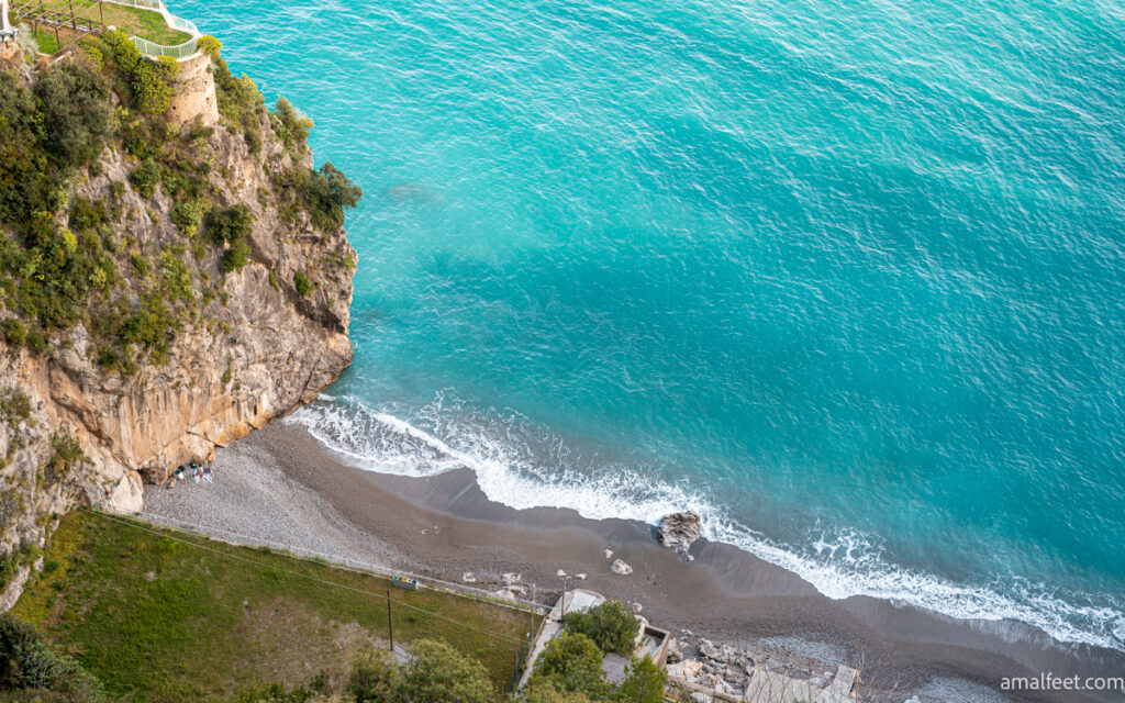The view of the Beach in the Castiglione area, under Ravello and near Atrani. Turquoise sea water, and vulcanic dark sandy shore, pictured from above. Rocky cliffs and greenery on the side.