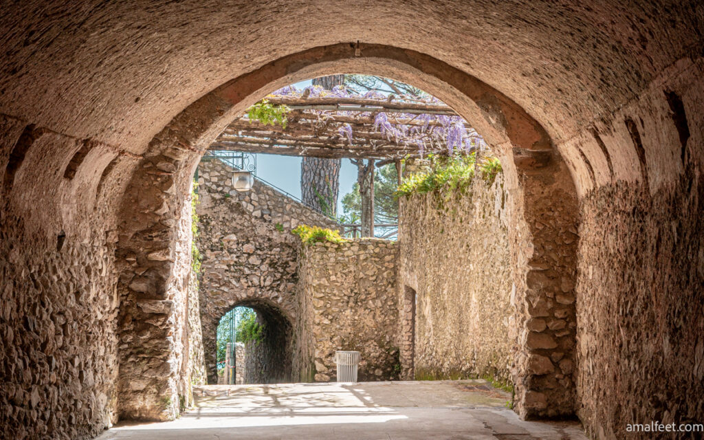 Arched alleyway in Ravello. Ancient looking stone walls. Purple wistera flowers hanging down from the Villa Rufolo's garden above.