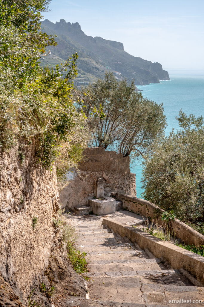 The footsteps between Minori and Ravello. Zig zagging and ancient looking stone staircase, and a drinking fountain in the turn. Trees, see and mountains in the background. 