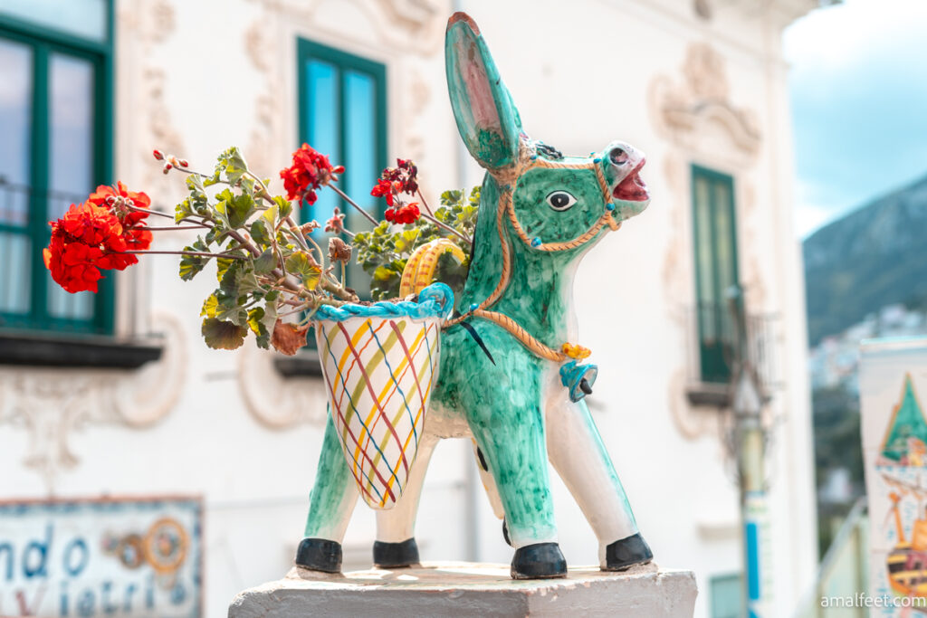 The little green donkey, made of Ceramic. Hand painted delighfull dunkey pottery in a top of a column. 
The donkey ffunctional as a flower pot, with red geranemum planted in the sack its carrying.