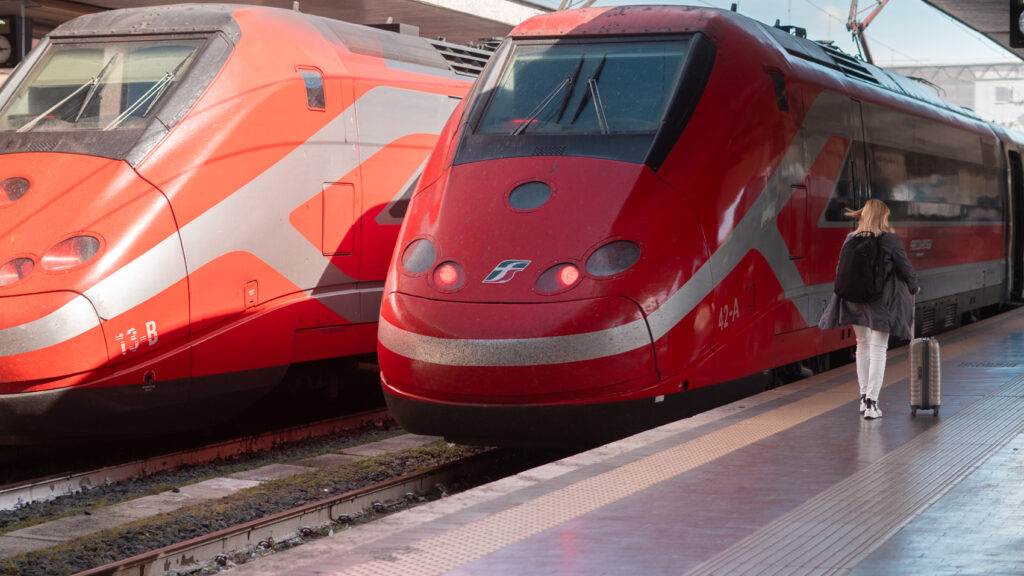 2 high-speed trains in the Central Station of Rome. Both train are red, with silver stripes. A women walks near the train on the platform with a small suitcase.