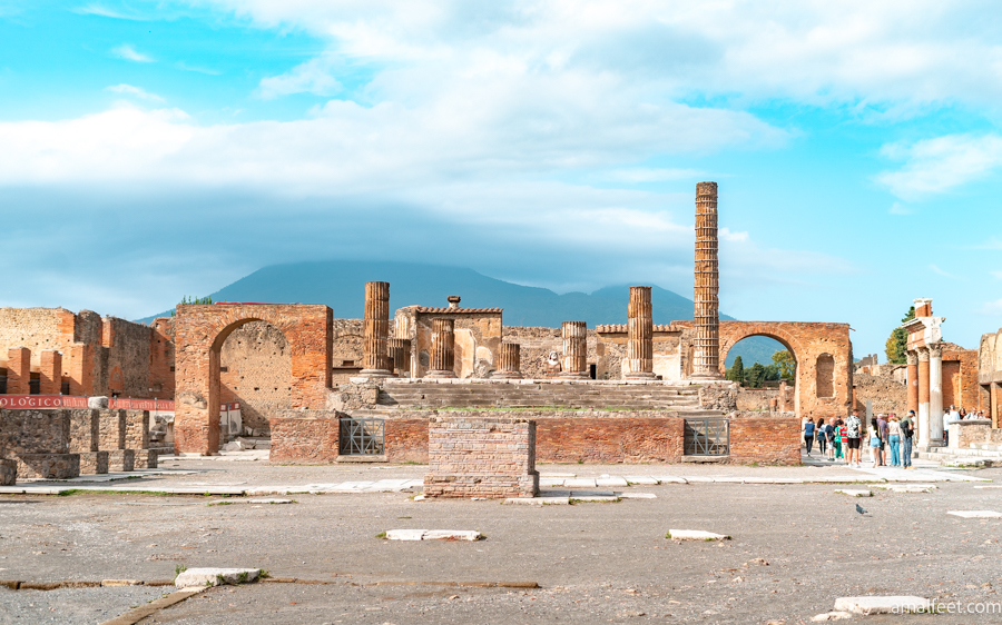 Pompeii, in a sunlit day. The ruins and the Vesuvius partially visible behind. The mountain top is covered with clouds.