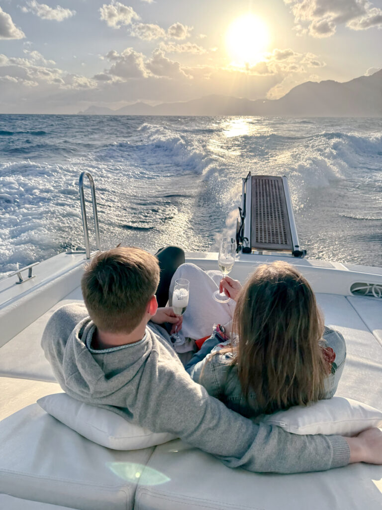 Couple relaxing on a boat, enjoying a sunset cruise with aperitivo. They are hugging each other with prosecco in hand, while watching the setting sun and the Amalfi Coast mountains silhouette in the background.